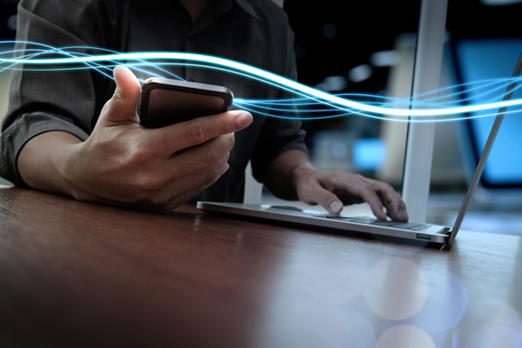Close-up of a person using a smartphone and laptop simultaneously, with digital light trails representing data connectivity.