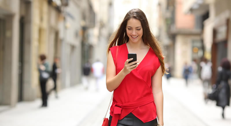 Young woman smiling while walking down a city street and looking at her smartphone, wearing a red top.